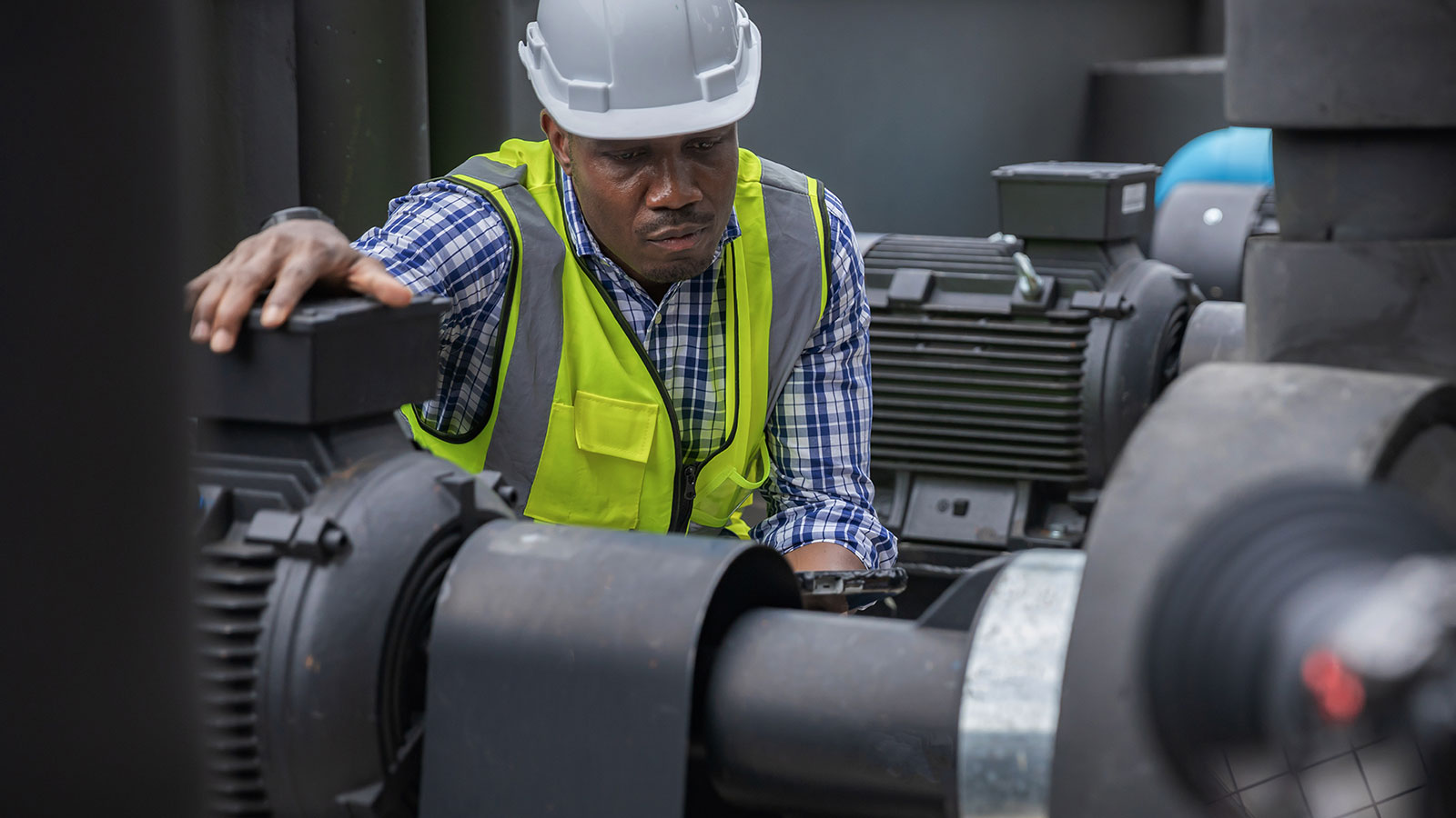 Maintenance engineer in a hard hat and safety vest inspecting an industrial pump motor in a manufacturing facility.